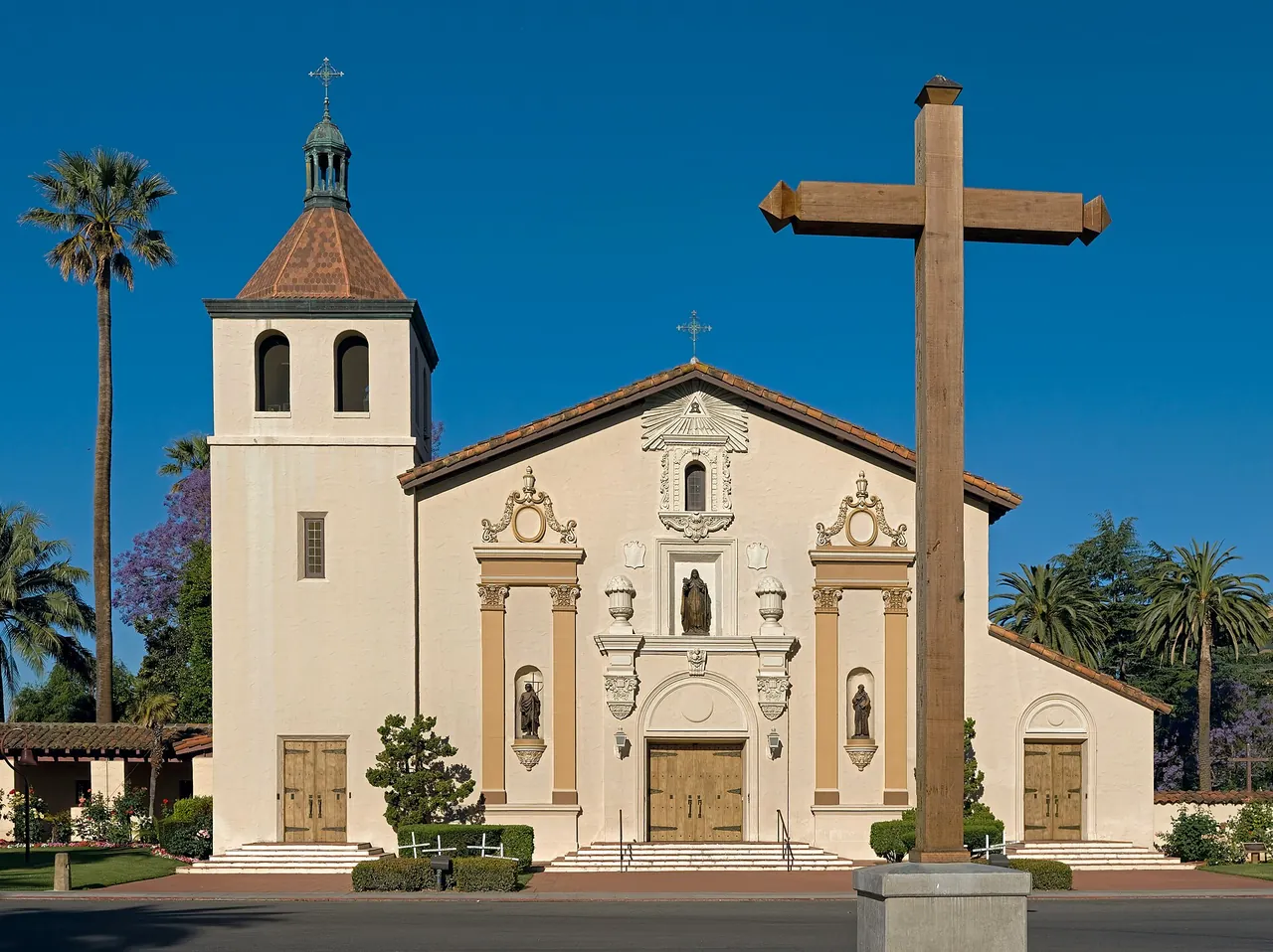 Mission Santa Clara de Asis with its bell tower and wooden cross under a clear blue sky.